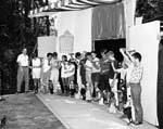Harry Burnett directs a group of children presenting the marionettes they have made at Idyllwild. They are standing on a puppet stage. 